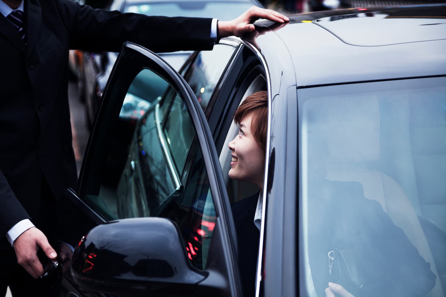 Man in a suit holding the rear door of a car open for a smiling woman in the back seat