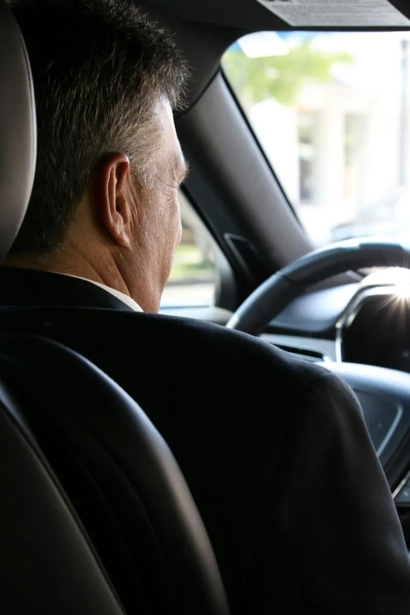 Man in a dark suit at the wheel of a car, seen from the back seat