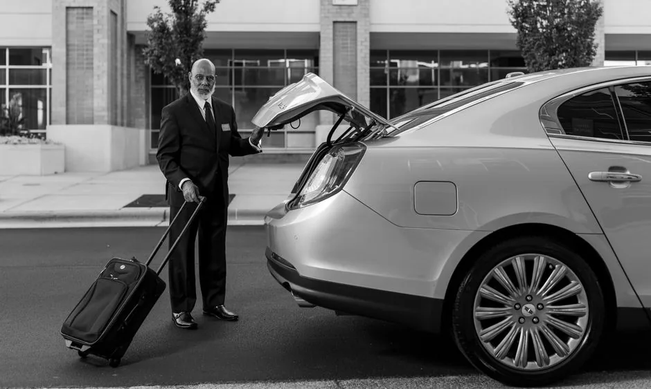 Chauffeur in a dark suit with a wheeled suitcase beside the open trunk of a sedan, in black and white