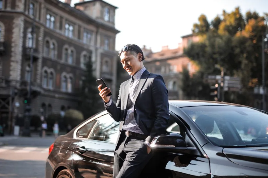 Man in a suit checking his phone beside a black sedan on a city street