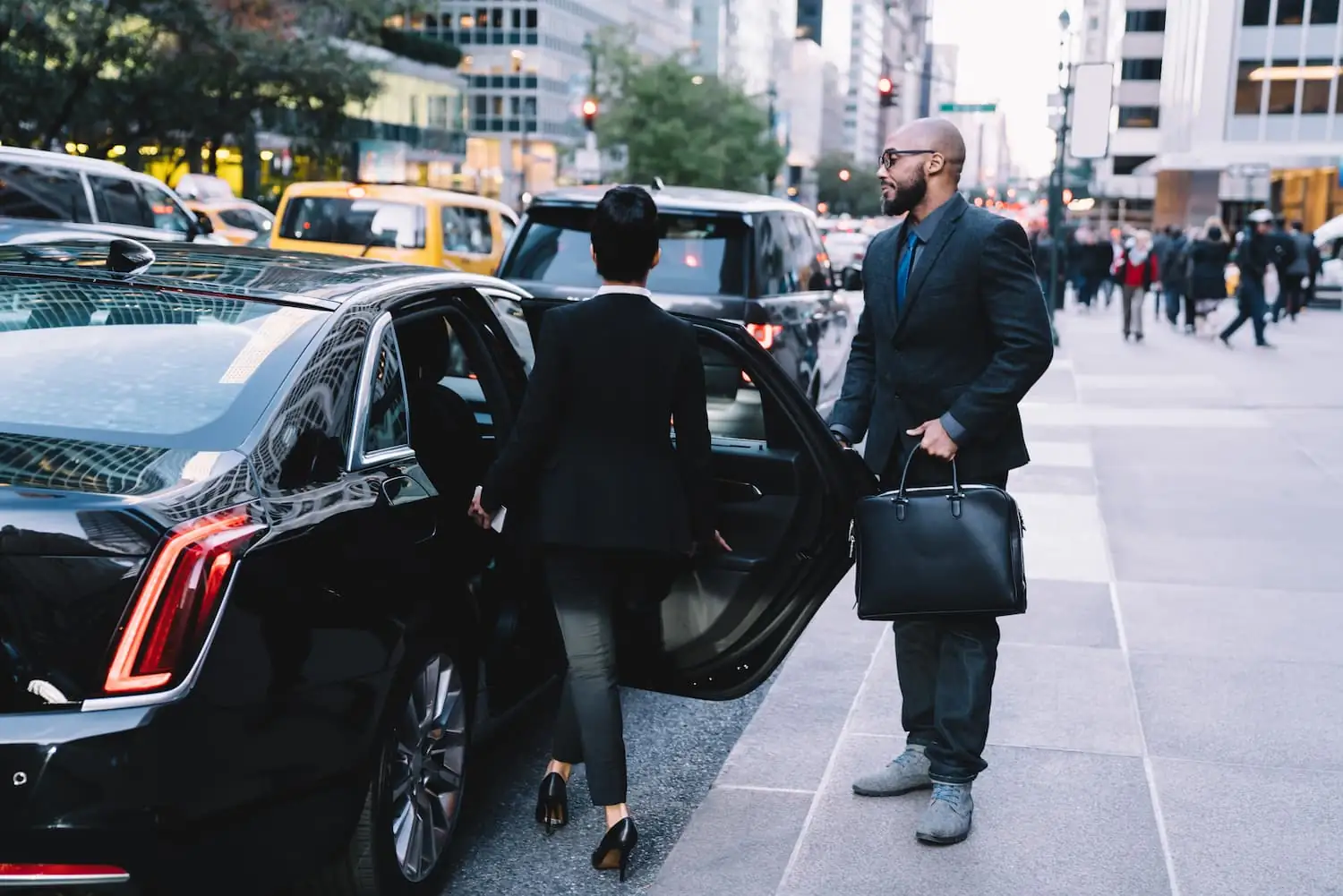 Chauffeur holding the rear door of a black sedan for a passenger on a busy city sidewalk with yellow cabs behind