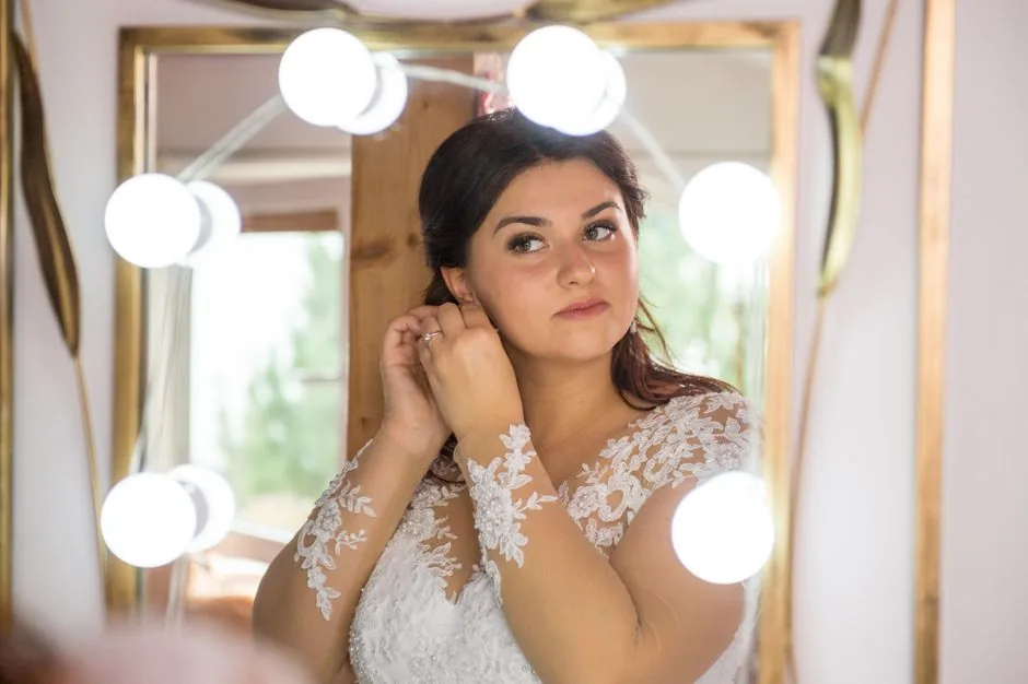 Bride fastening an earring in front of a lit mirror